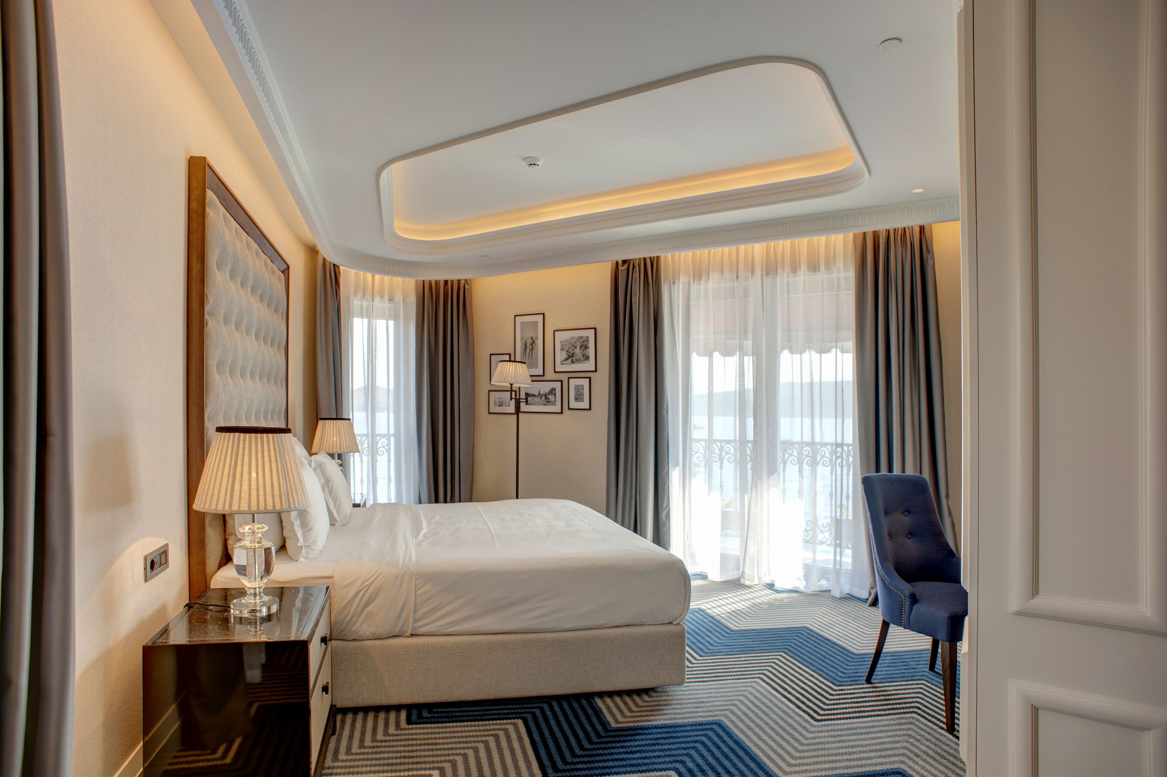 A hotel room with a large bed, a desk with a lamp, a chair, and a view of the outdoors through the windows.