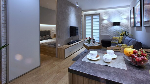 A modern living room with a sofa, coffee table, and a TV. There is a bed in the background and a window with blinds. - 4UApart - Apartment suite Picasso (Swinoujscie)
