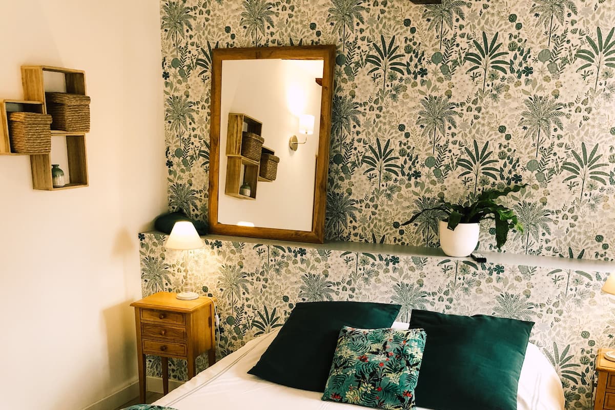 A bedroom with a floral-patterned wallpaper, a bed with a green floral bedspread, two pillows, a nightstand with a lamp, and a framed mirror on the wall.