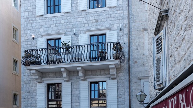 Un immeuble en pierre à plusieurs étages, doté de volets blancs, d’un balcon avec des balustrades en fer forgé et d’une entrée au niveau du sol avec une porte noire.