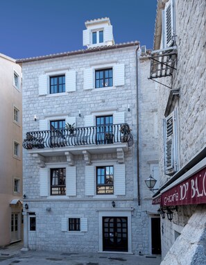 A multi-story stone building with white shutters, a balcony with wrought iron railings, and a ground-level entrance with a black door. - Villa Luna (Trogir)