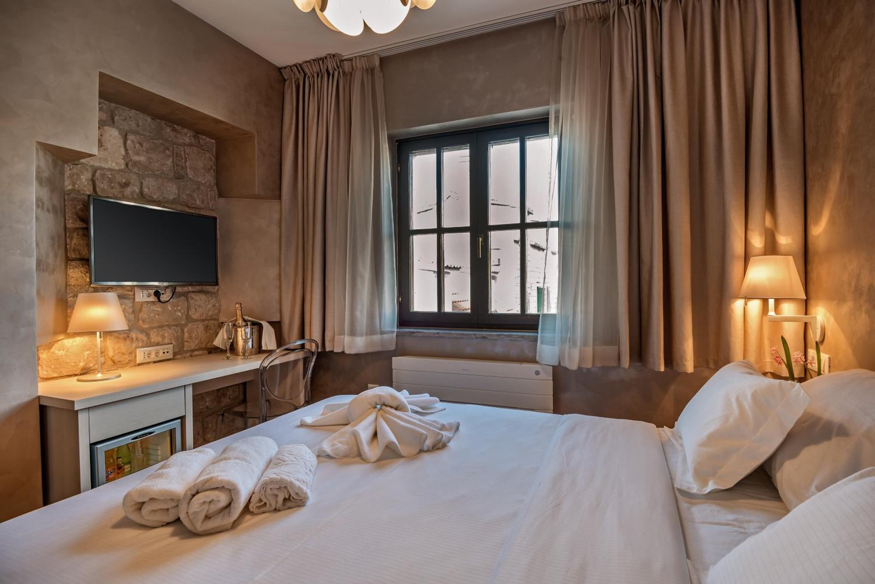 A hotel room with a bed, a flat-screen TV mounted on a stone wall, a desk with a lamp, and a window with curtains.