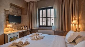 A hotel room with a bed, a flat-screen TV mounted on a stone wall, a desk with a lamp, and a window with curtains.