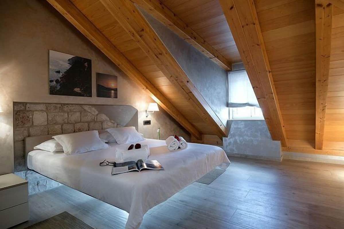 A bedroom with a stone headboard, wooden ceiling beams, a bed with white linens, a nightstand, and a window.