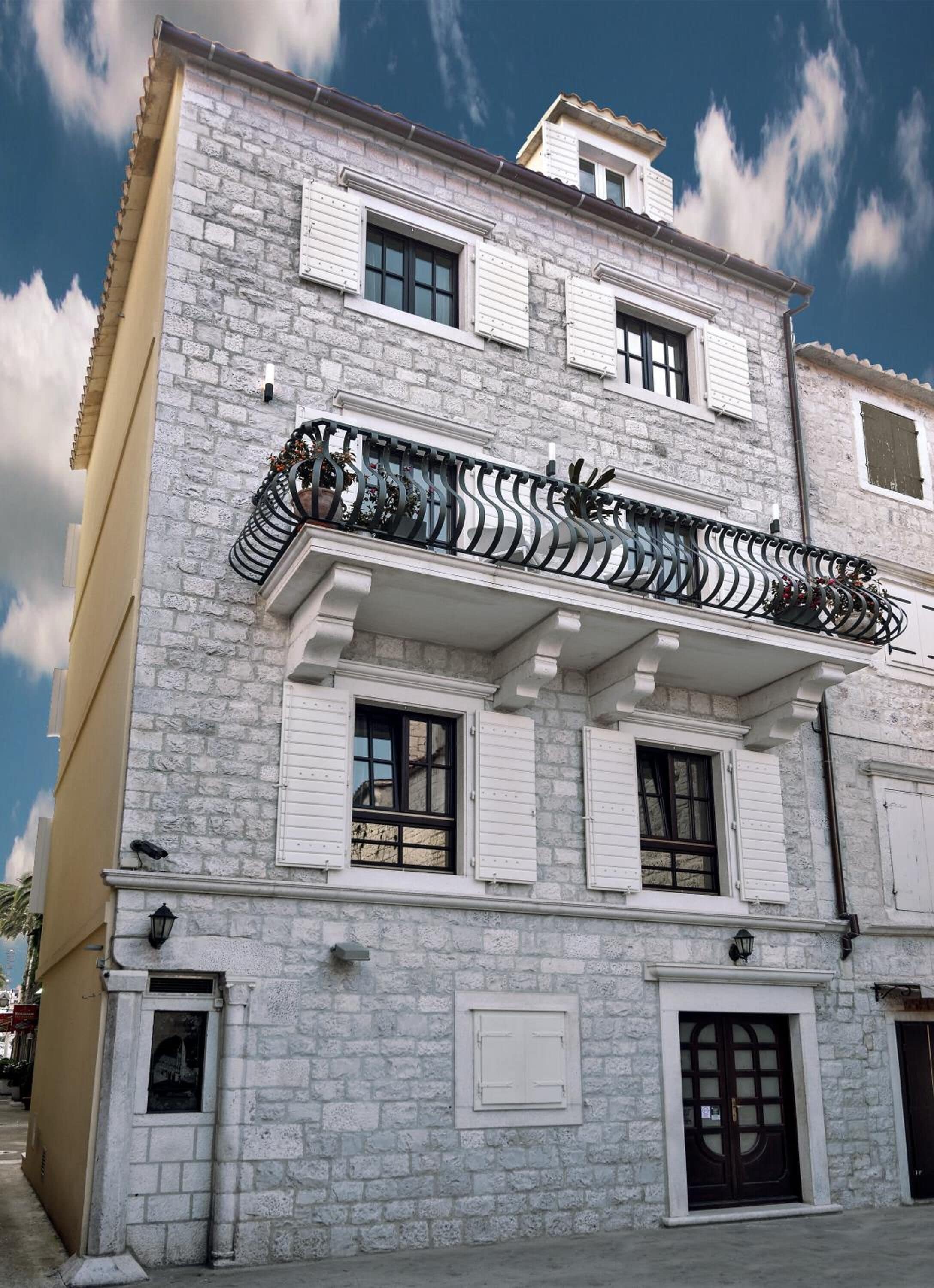 A two-story building with a stone facade, white shutters, and a balcony with wrought iron railings.