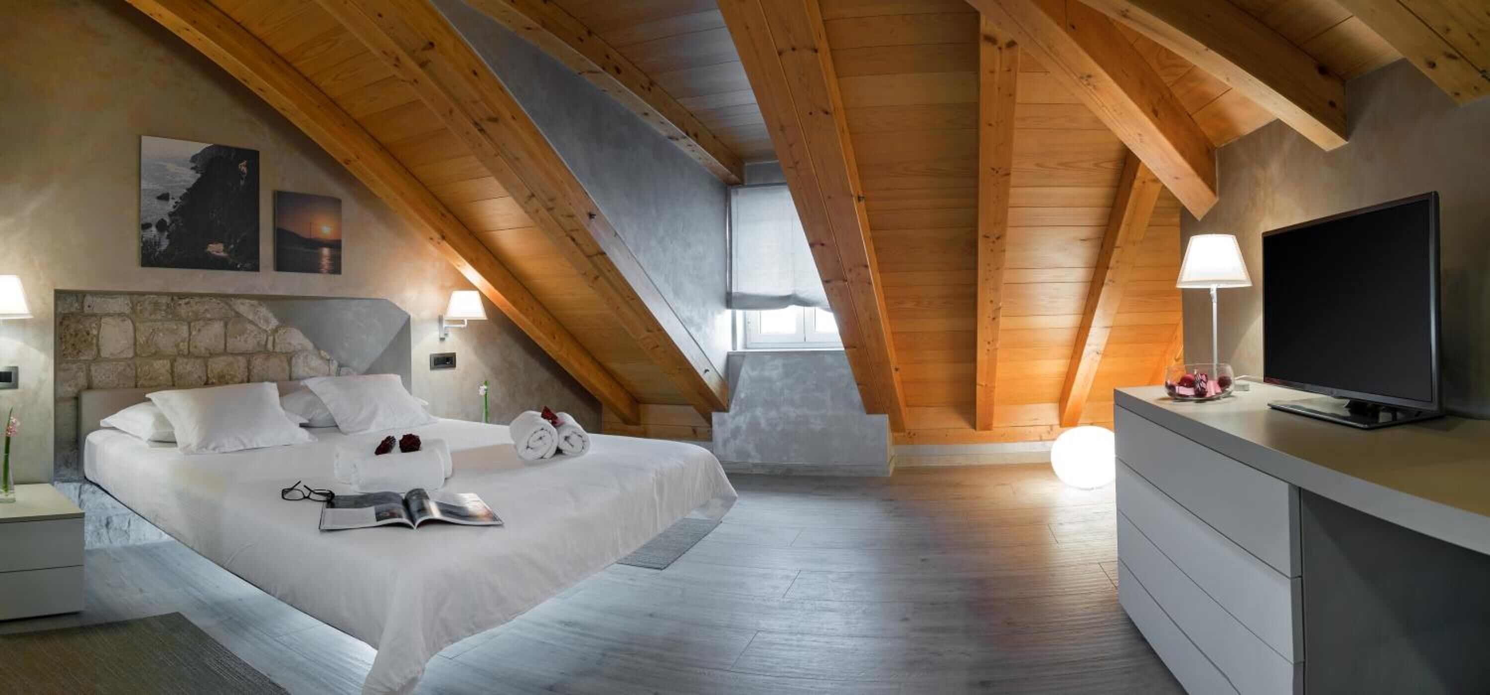 A bedroom with a sloped ceiling, a bed with white linens, a stone wall, a television, and a dresser.