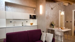 Une cuisine moderne avec des meubles blancs, un canapé violet et un coin repas avec des chaises transparentes et une table blanche.