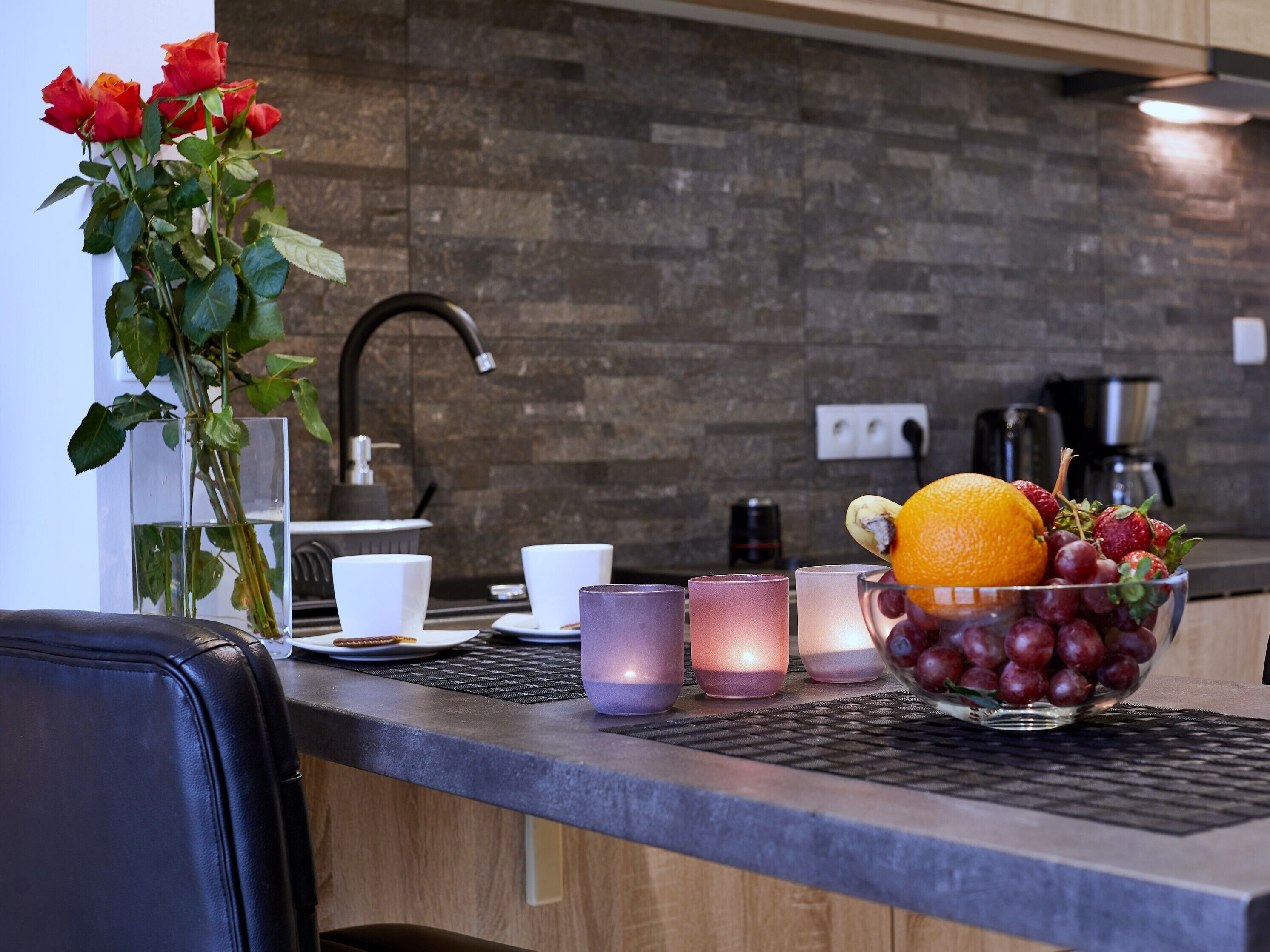 Eine Küchenarbeitsplatte mit einer Obstschale, die Weintrauben, eine Orange und Erdbeeren enthält, einer Rosenvase und einer Kaffeemaschine.