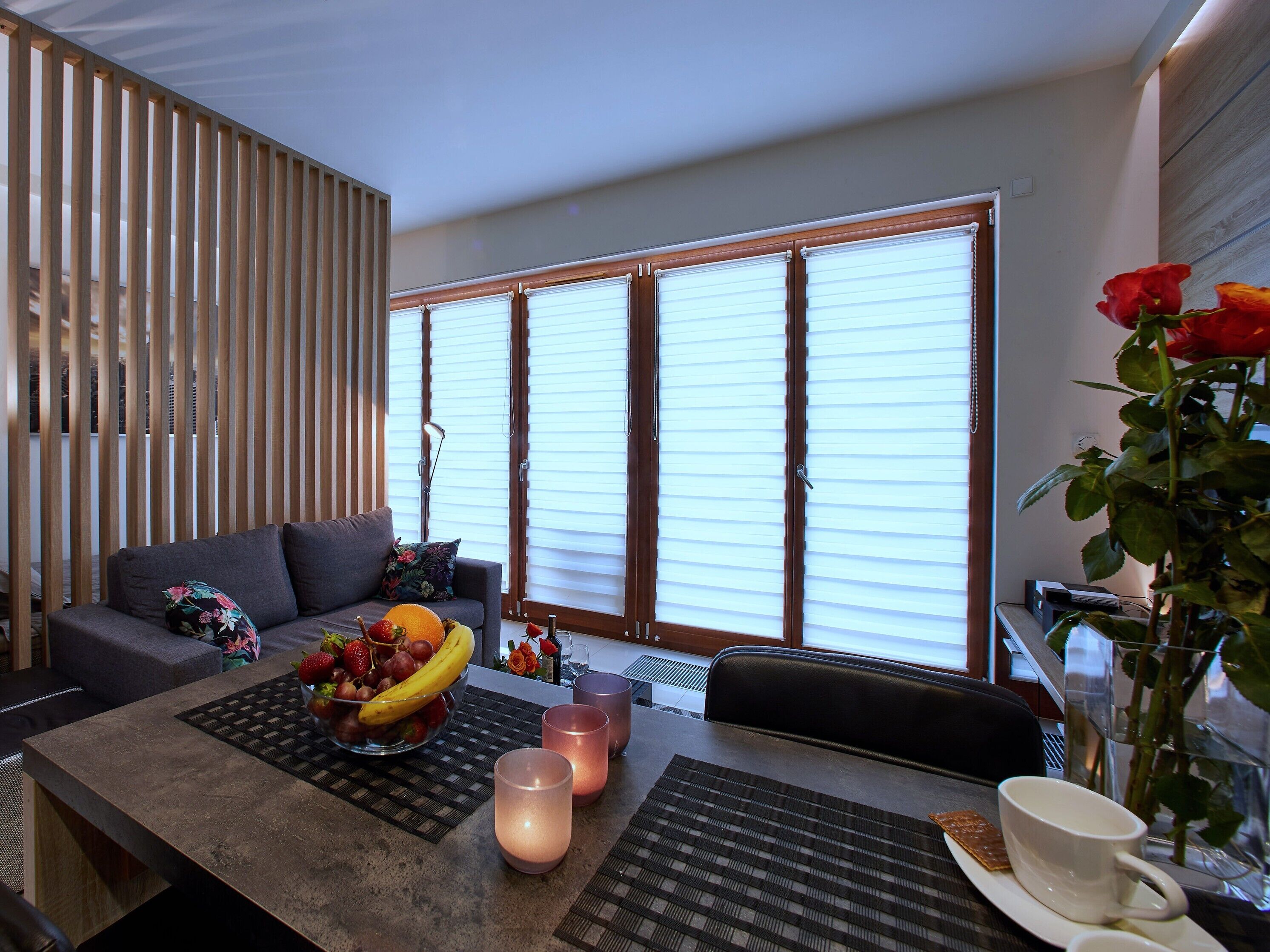 A modern living room with a sofa, a dining table with candles, a fruit bowl, and a vase with flowers.