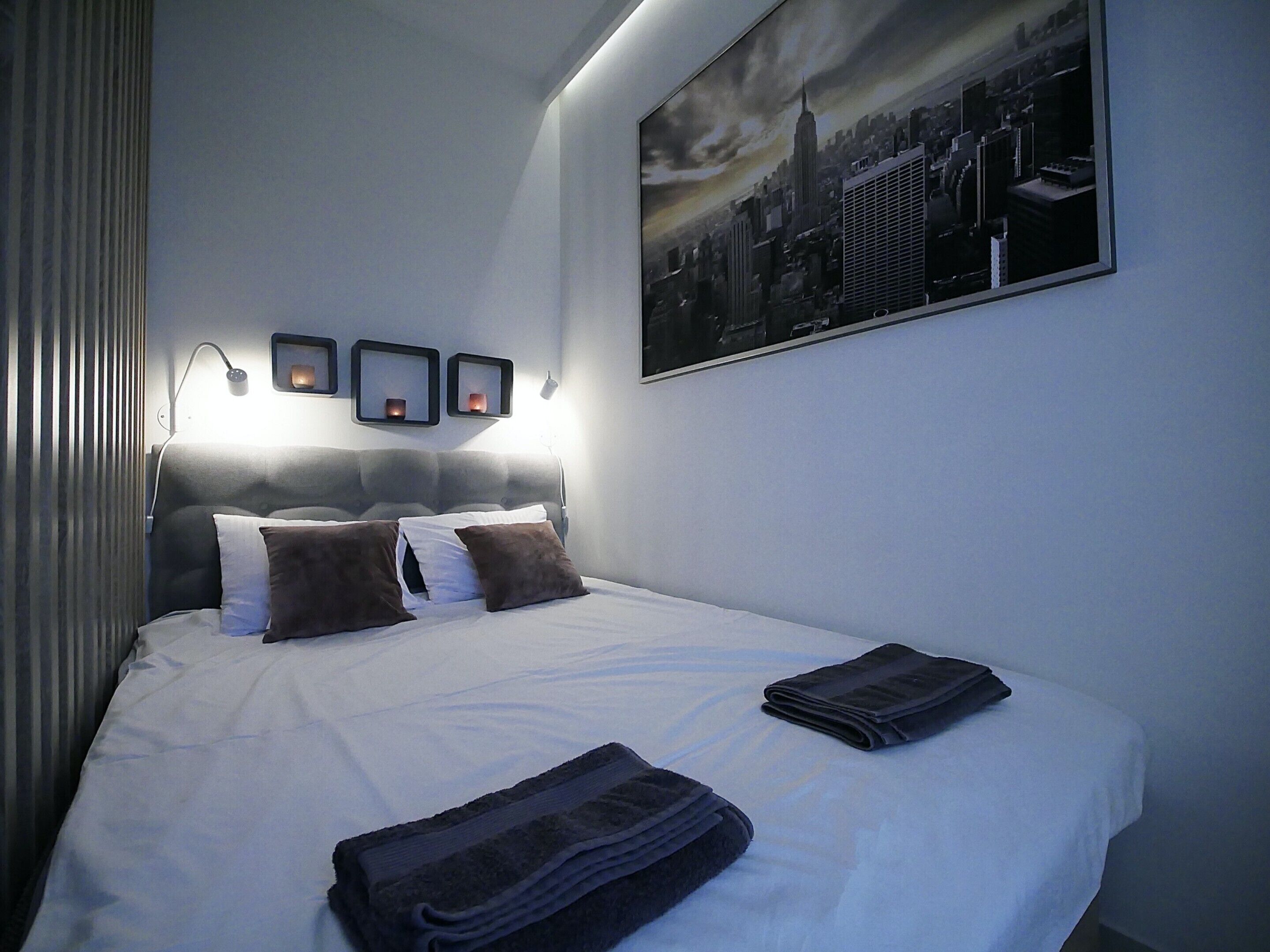 Una cama bien hecha con almohadas, una toalla doblada y un cuadro con una vista urbana en la pared.