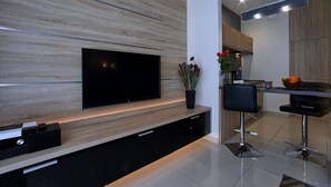 A modern kitchen with a flat-screen TV mounted on a wooden wall, a dining area with a black chair, and a kitchen island with a bowl of fruit. - 4UApart - Apartment studio Platan New York (Swinoujscie)