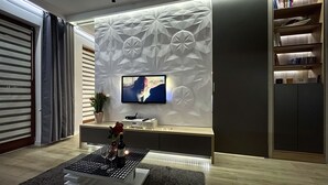 Una sala de estar moderna con un televisor de pantalla plana montado en una pared con textura, una mesita auxiliar con un florero y copas de vino, y una estantería a la derecha.