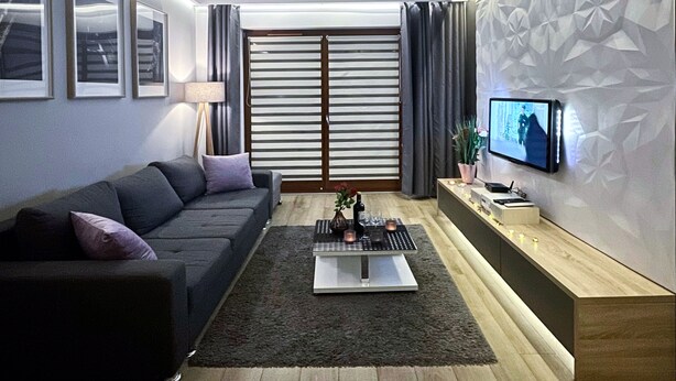 Una sala de estar moderna con un sofá gris oscuro, una mesita de centro con velas, un televisor en la pared y un papel tapiz con un estampado geométrico.