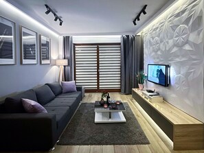 Ein modernes Wohnzimmer mit einem dunkelgrauen Sofa, einem Couchtisch mit Kerzen, einem an der Wand befestigten Fernseher und einer Wand mit geometrischem Muster.