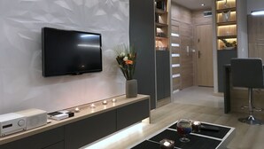 Una sala moderna con un televisor de pantalla plana, una mesa consola con dispositivos electrónicos, un florero con flores y una zona de bar con una copa y un control remoto.