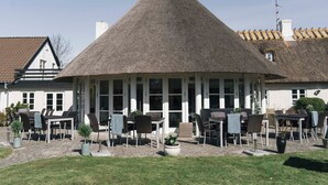 A building with a thatched roof and a covered outdoor seating area with tables and chairs. - Havgaarden Badehotel (Vejby)