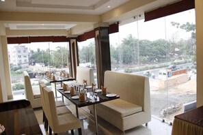 A dining area with tables set for four, a sofa, and a view of the cityscape. - Regenta Inn Larica (Barasat)