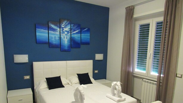 Ein Hotelzimmer mit einem großen Bett, weißer Bettwäsche, einem Kopfteil, zwei Nachttischen, einem Fenster mit Jalousien und einer Wand mit einem blauen Gemälde.