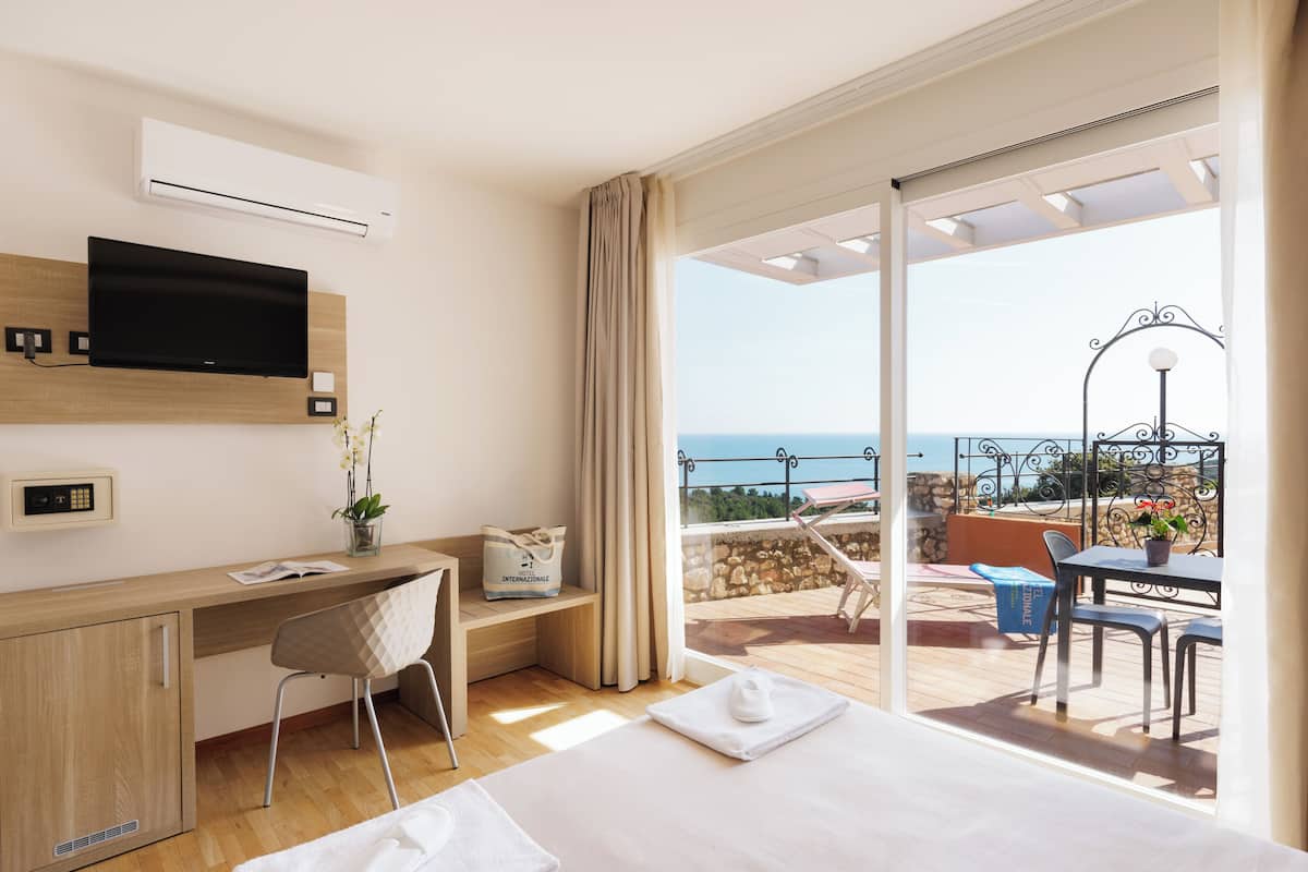 A hotel room with a bed, a desk, a TV, a balcony with a view of the sea, and a dining area.