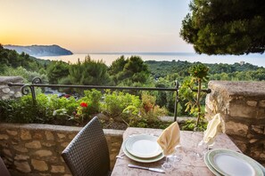 A table set for two with plates, cutlery, and glasses, overlooking a scenic view of a coastline with trees and a distant mountain.