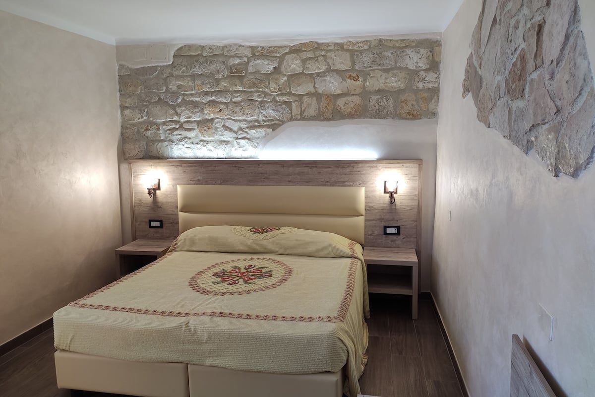 A bedroom with a stone wall, a bed with a floral bedspread, bedside lamps, and a nightstand.