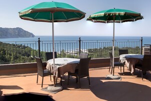 A terrace with a table and chairs, two green umbrellas, and a view of the sea and hills.