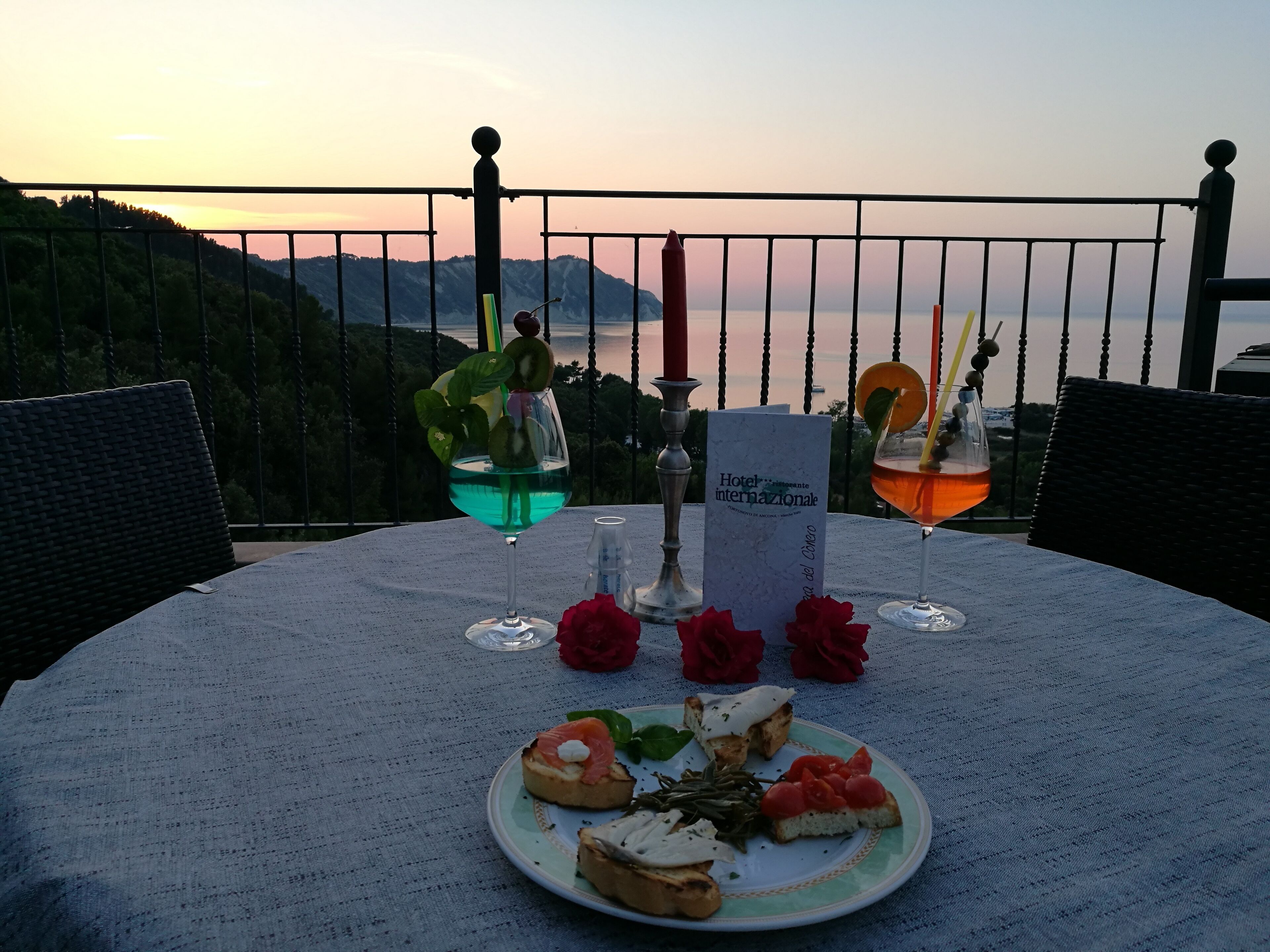 A table set with a plate of appetizers, two drinks, a menu, and decorative roses on a balcony overlooking a scenic view.