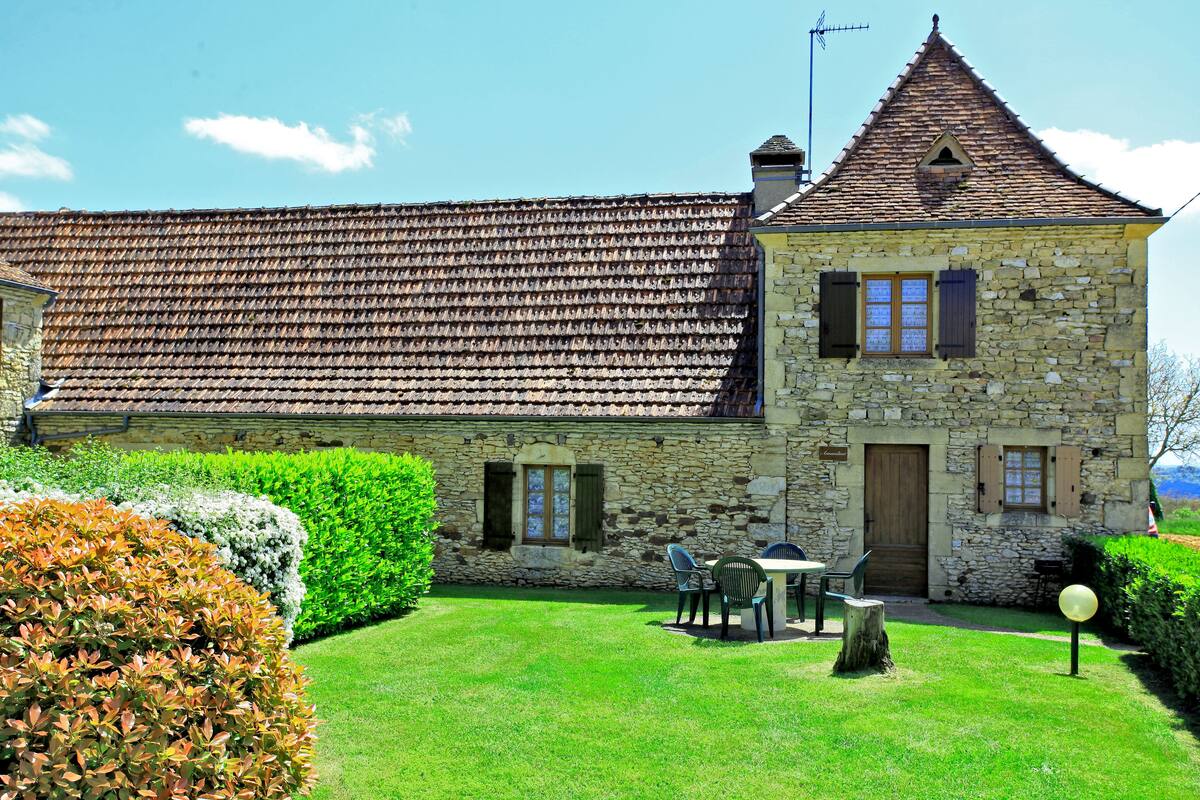 Un bâtiment en pierre avec un toit en tuiles, un petit jardin avec une table et des chaises, et une porte en bois.