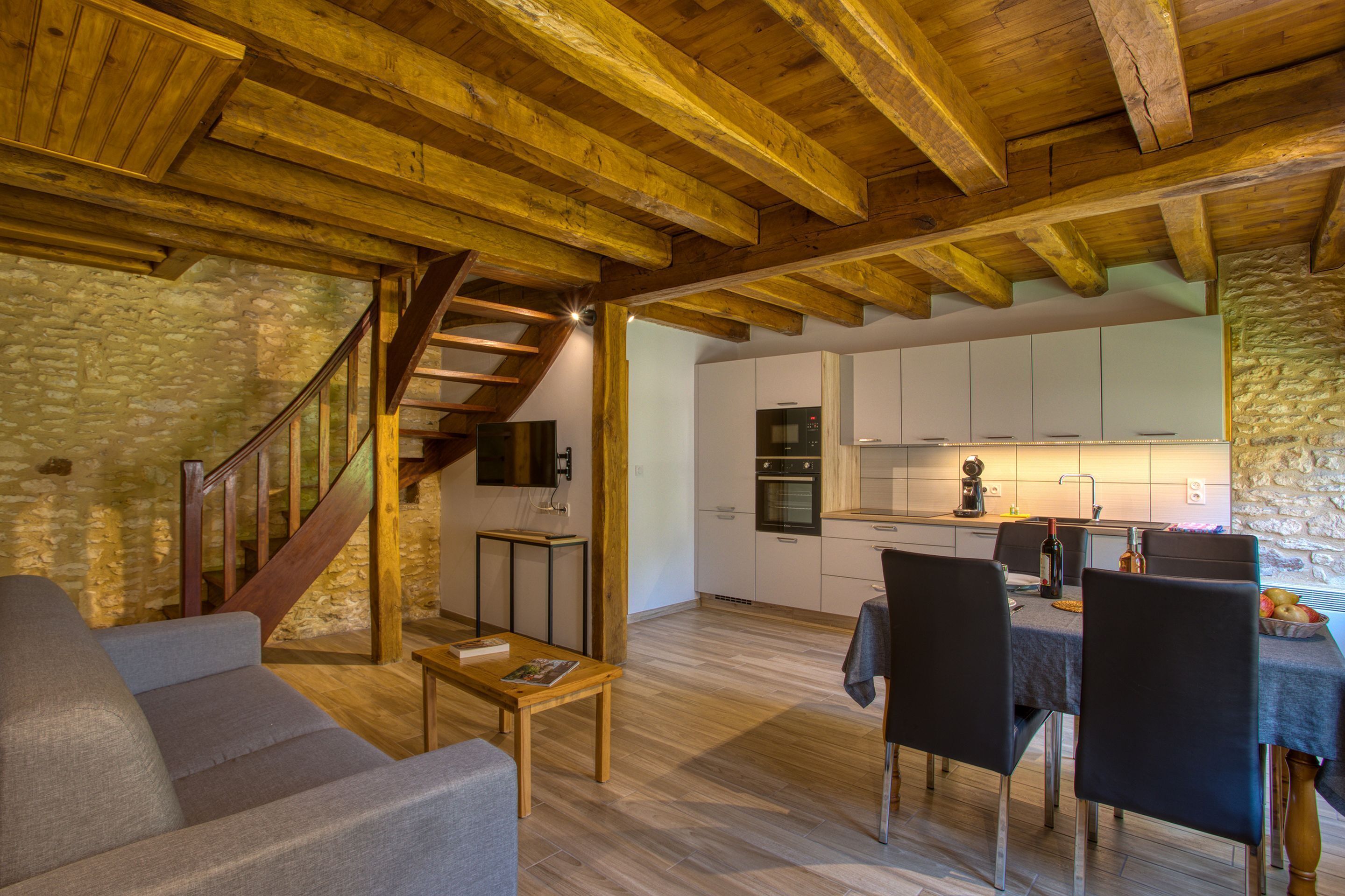 A spacious living area with a wooden ceiling, a stone wall, a staircase, a kitchen, and a dining area with chairs and a table.