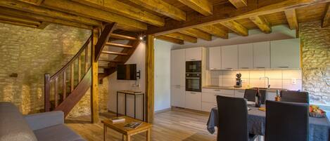A spacious living area with a wooden ceiling, a stone wall, a staircase, a kitchen, and a dining area with chairs and a table.
