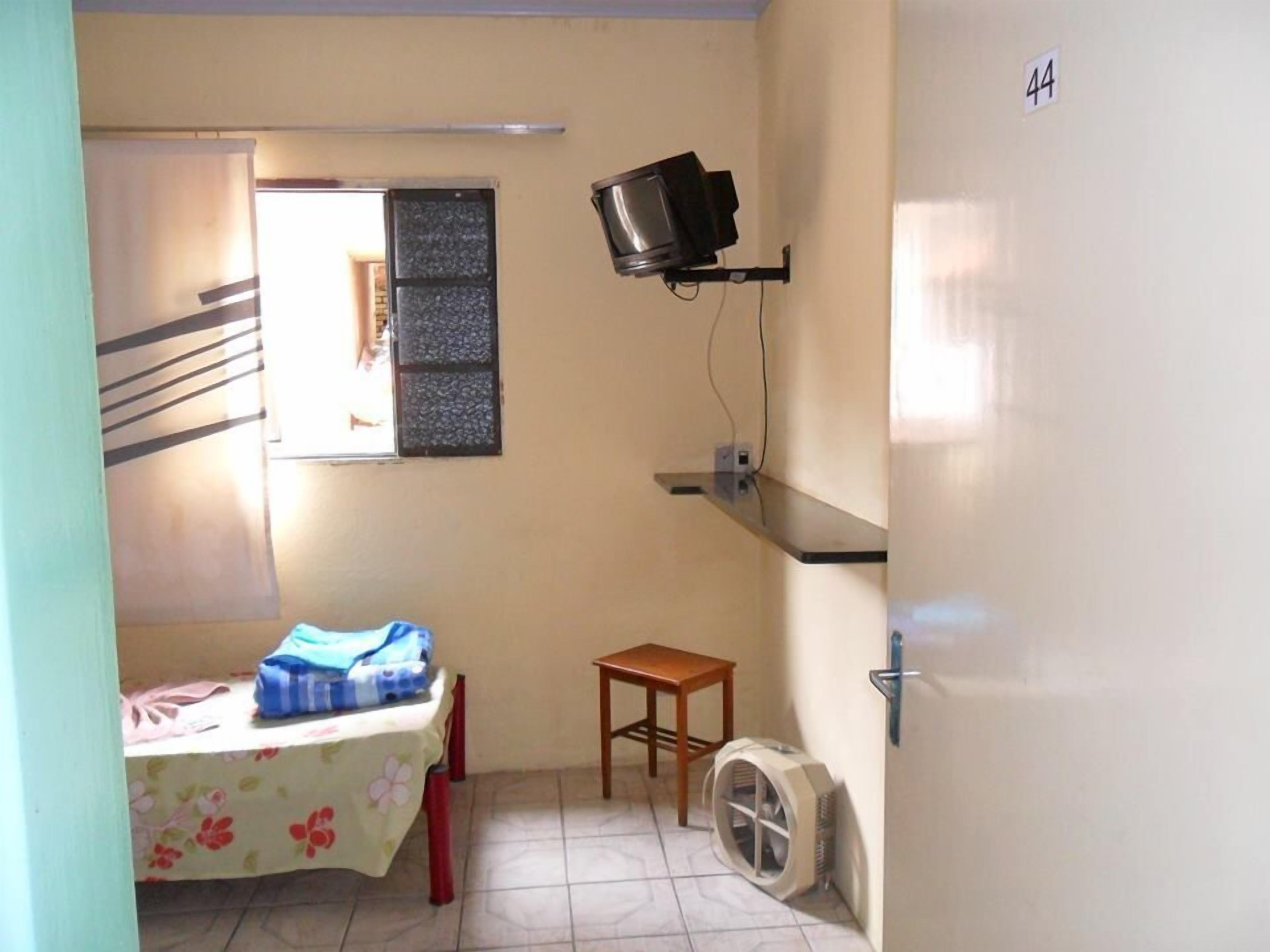 Quarto pequeno com cama, mesa, TV fixada na parede, janela e porta.