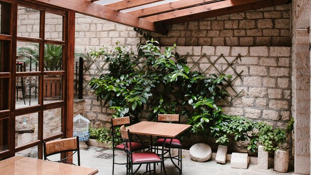 Un patio techado con techo de madera, piso de piedra y pared de ladrillo. Hay mesas y sillas de madera, y plantas en macetas.