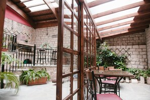Un patio interior con mesa y sillas de madera, rodeado de plantas en macetas y un muro de piedra.
