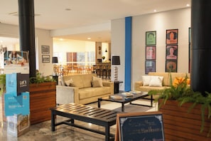 A hotel lobby with a sofa, chairs, a coffee table, and a dining area in the background. - Transamerica Fit Jaú (Jau)