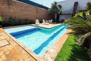 A rectangular swimming pool with clear blue water, surrounded by a brick wall, a grassy area, and some plants. - Transamerica Fit Jaú (Jau)