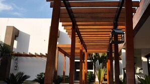 Un paseo techado con vigas de madera y suelo reflectante, que conduce a un edificio de diseño moderno bajo un cielo despejado.