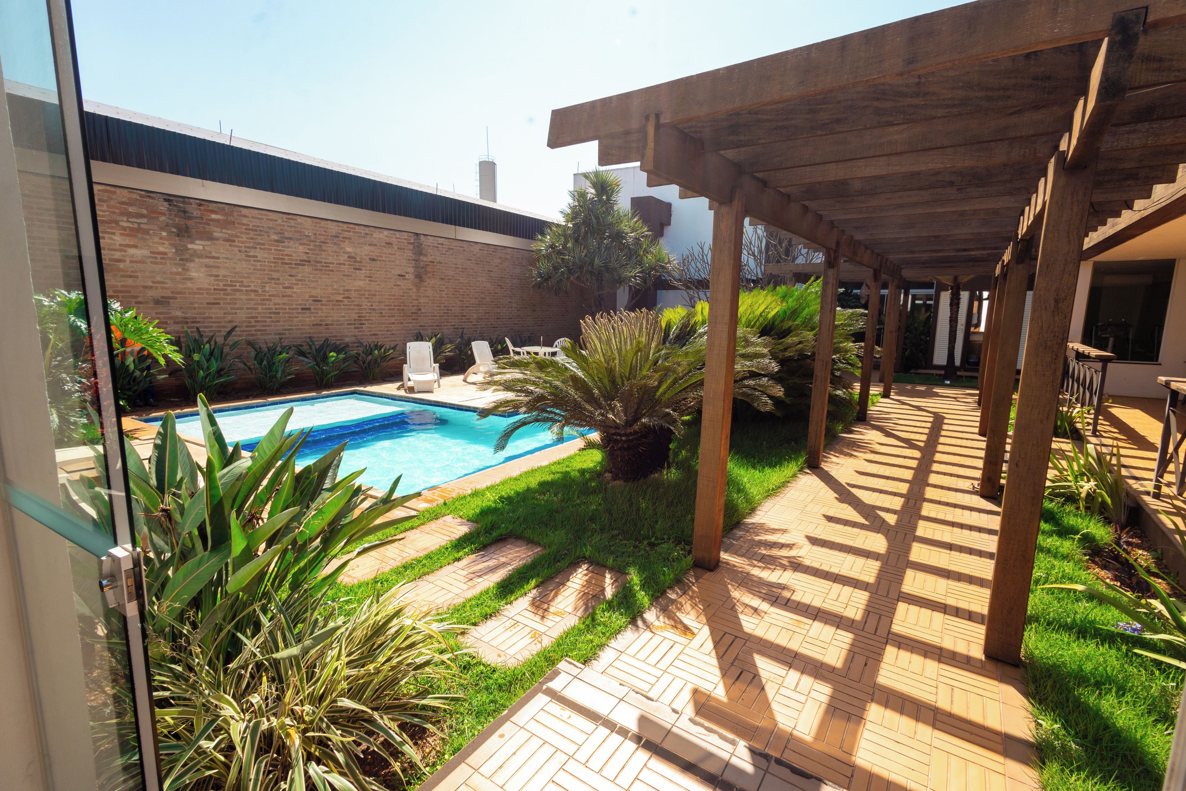 A pool area with a wooden pergola, a brick wall, and various plants.