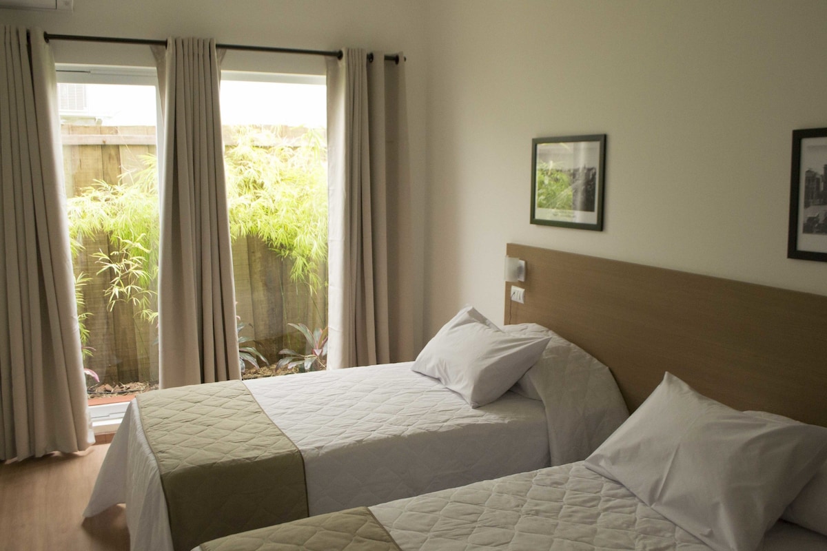 A hotel room with two beds, a view of greenery outside, and a framed picture on the wall.