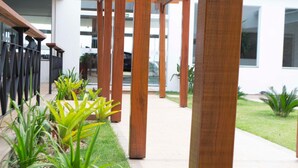 Un espacio al aire libre cubierto con columnas de madera, un sendero empedrado y diversas plantas verdes.