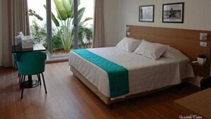 Habitación de hotel con cama, escritorio con silla, ventana con cortinas y una planta.