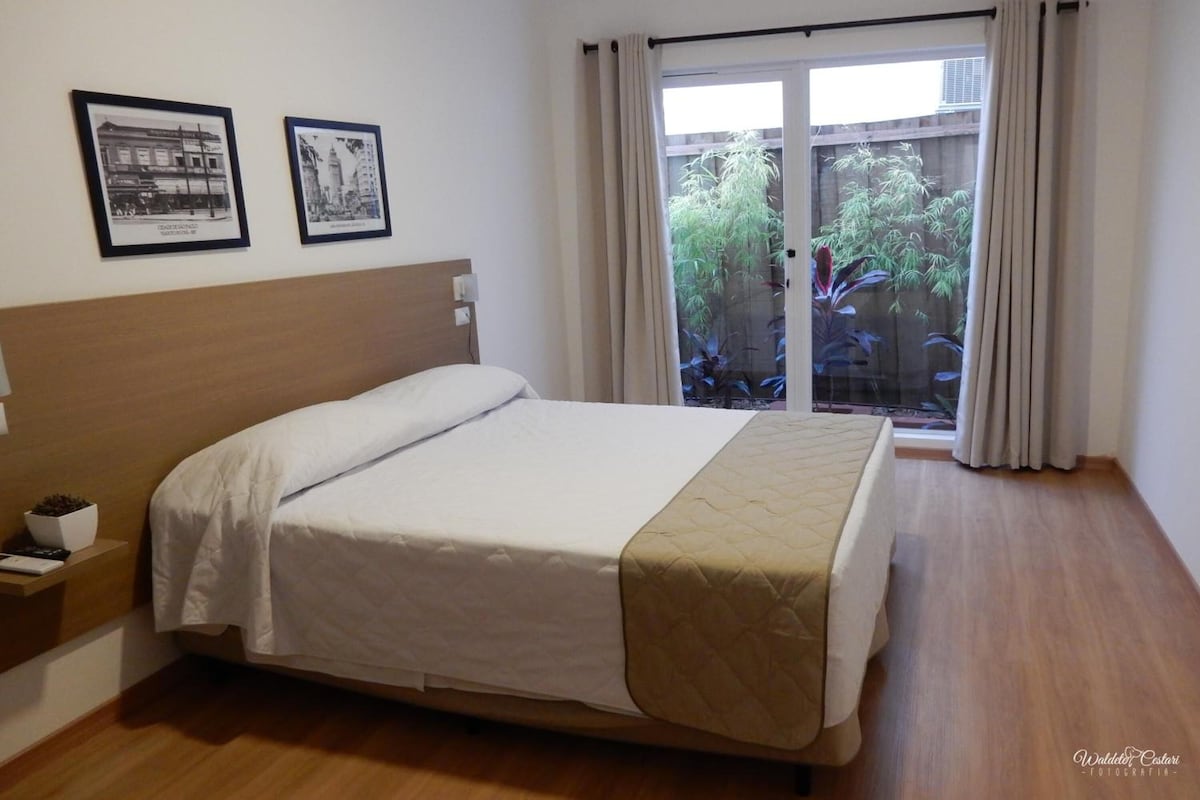 Quarto de hotel com uma cama, uma mesa de cabeceira, uma janela com cortinas e duas molduras na parede.