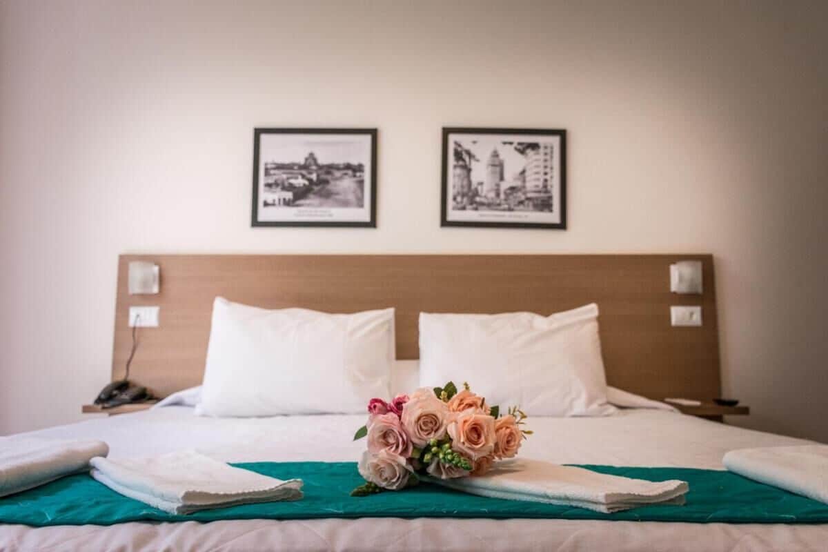 Uma cama bem arrumada com lençóis brancos, um buquê de rosas cor-de-rosa e duas fotografias preto e branco de paisagens urbanas na parede.