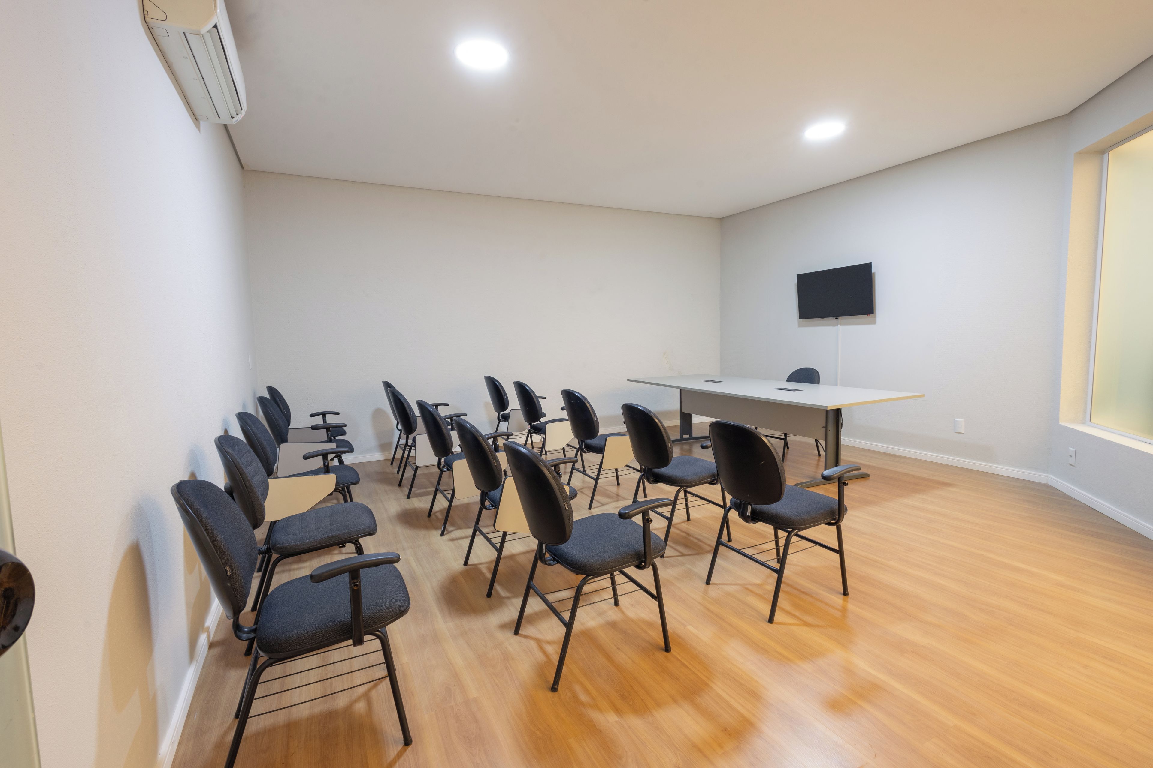 A conference room with a long table, multiple chairs, a flat-screen TV mounted on the wall, and wooden flooring.