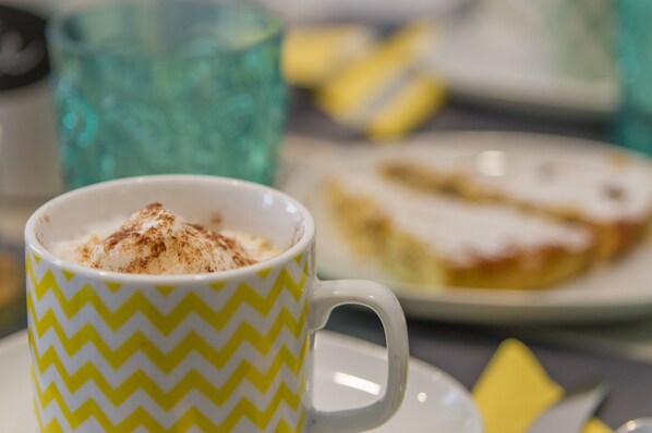 Una taza de café con canela espolvoreada encima, una porción de pastel en un plato y un vaso azul al fondo.