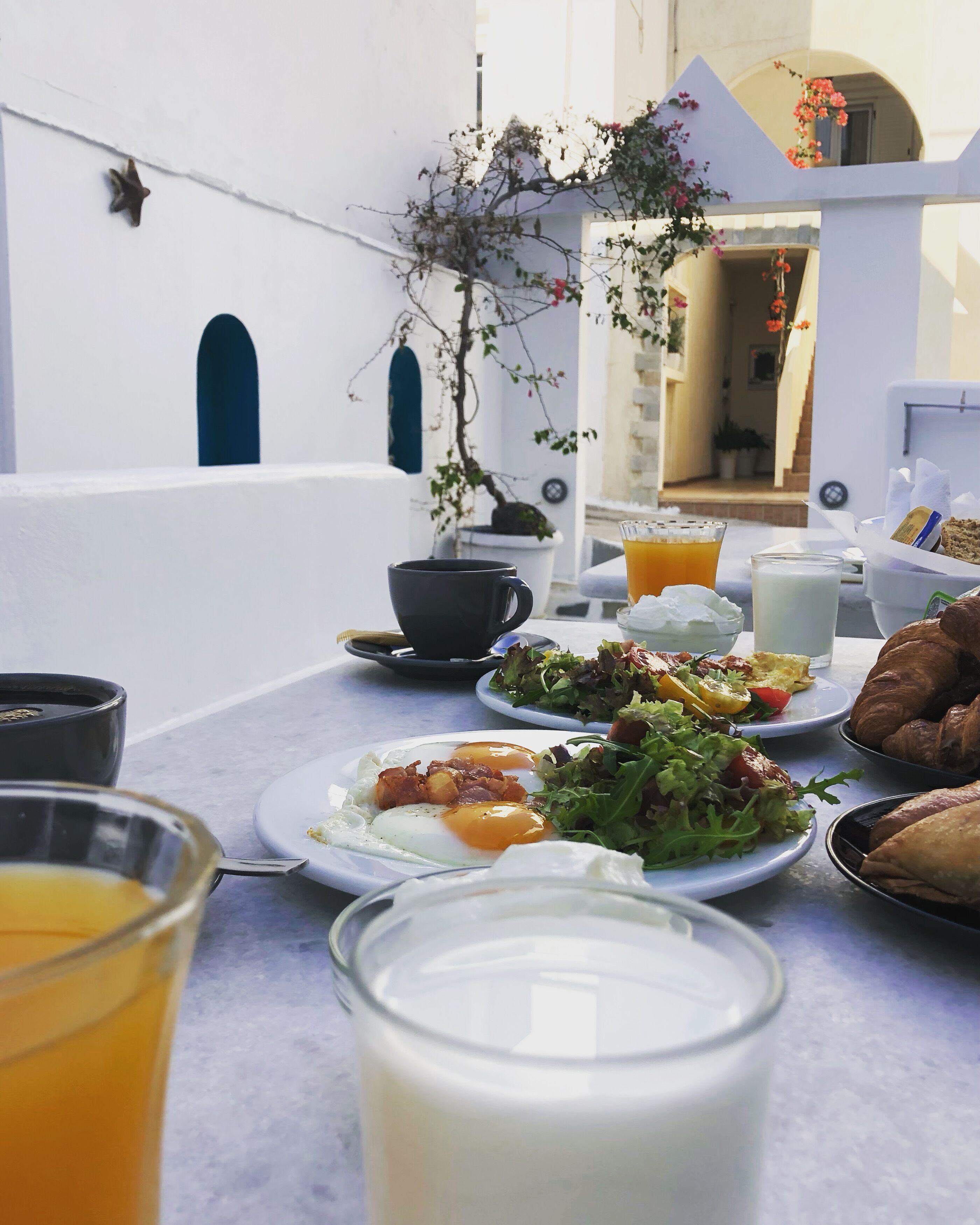 A table set with breakfast items including eggs, toast, and a salad, with a potted plant and a view of a building in the background.