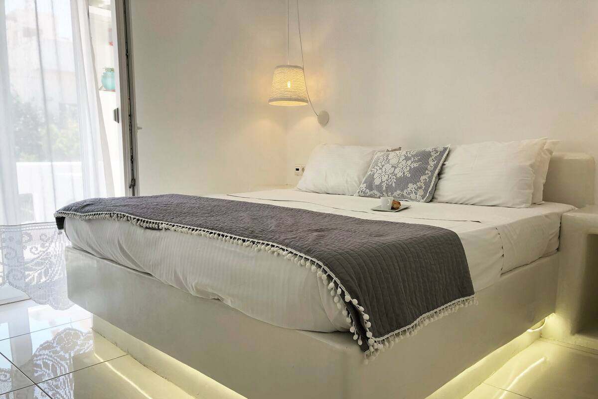A modern bedroom with a large bed, white bedding, a grey throw blanket, and a wall-mounted light fixture.