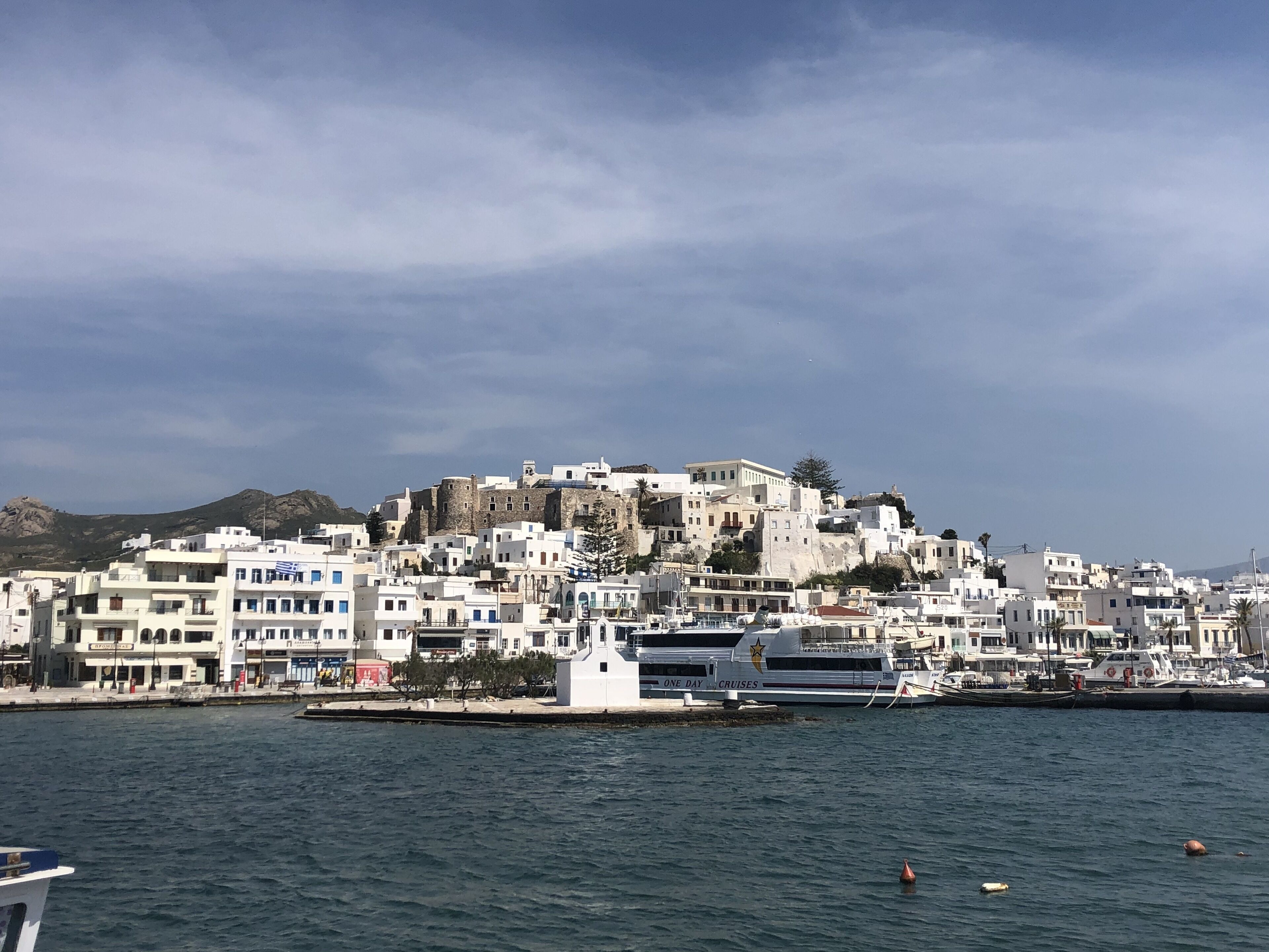 A coastal town with white buildings, a prominent hilltop structure, and a marina with boats.