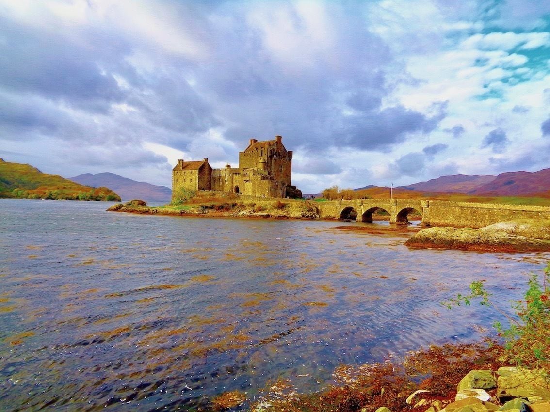 A historic castle by a river with a bridge leading to it.