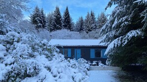 A snow-covered landscape with a house featuring a blue door and windows, surrounded by snow-laden trees and bushes. - Loch Ness Highland Cottages (Inverness)