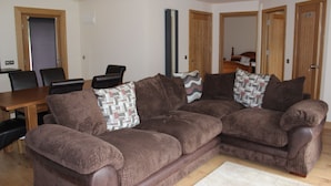 A living room with a brown sectional sofa, a wooden dining table, and a patterned rug. - Loch Ness Highland Cottages (Inverness)